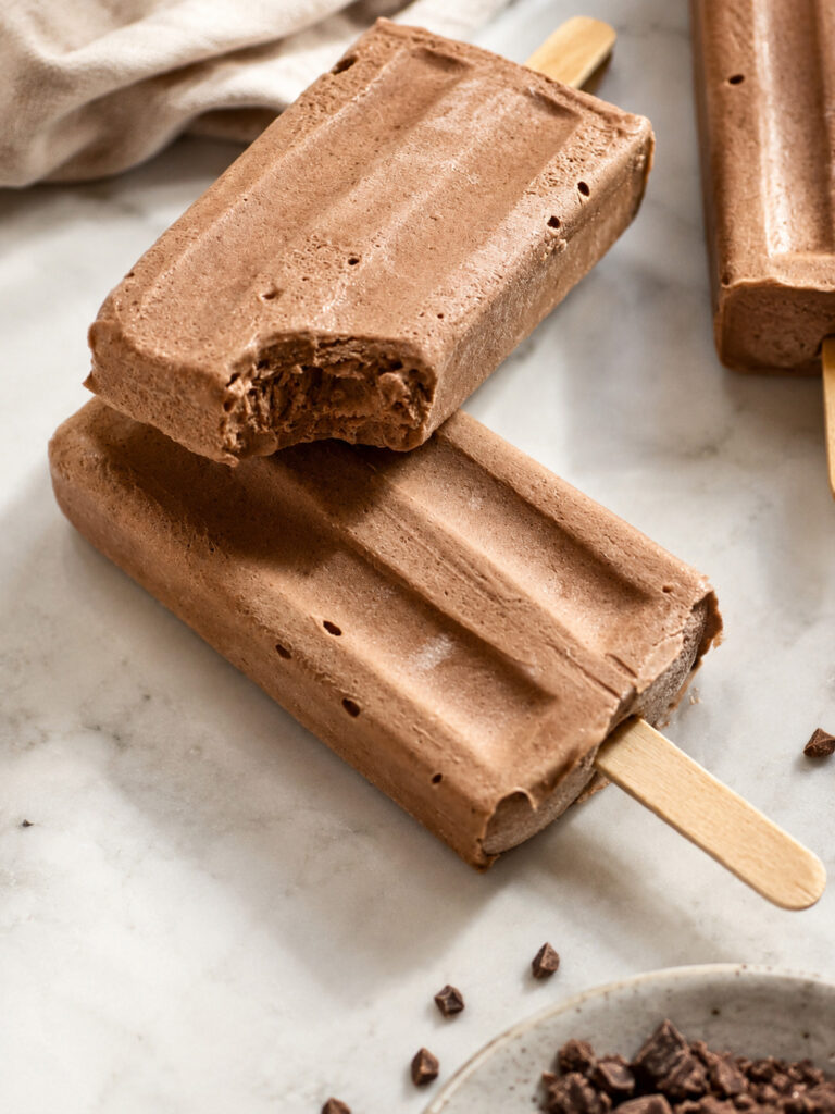 Close-up of creamy homemade chocolate popsicles with a bite taken out, showing the smooth texture on a marble surface with chocolate crumbs.