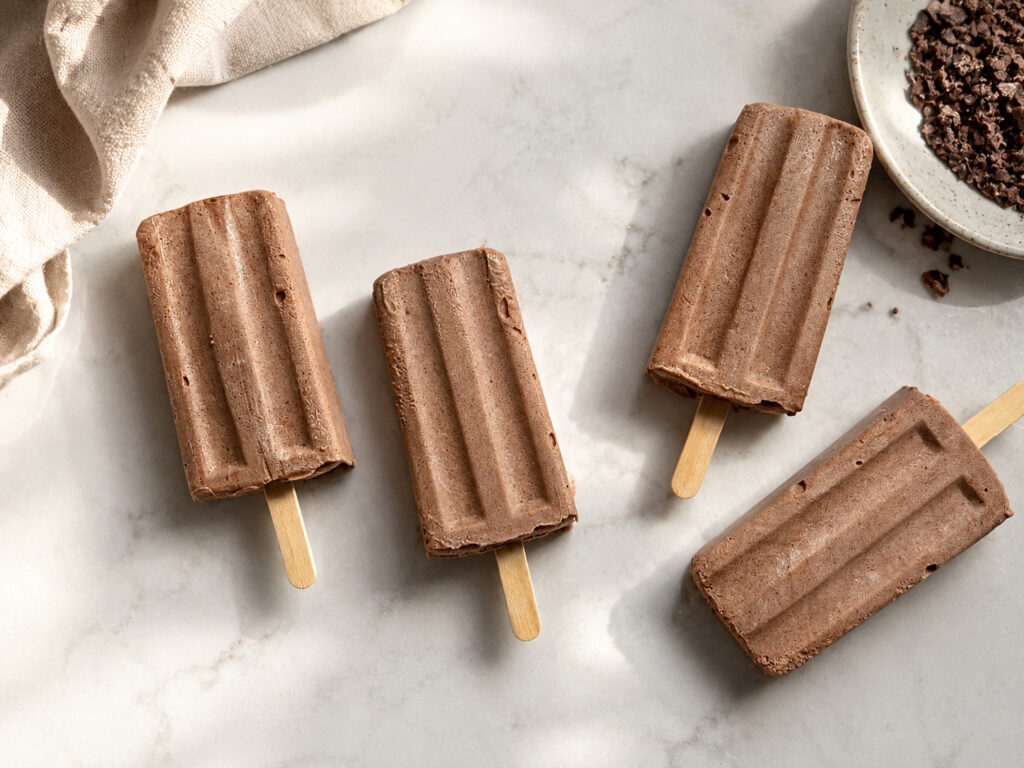 Four homemade chocolate popsicles arranged on a marble background with chocolate pieces and a neutral linen towel for a summer dessert recipe.