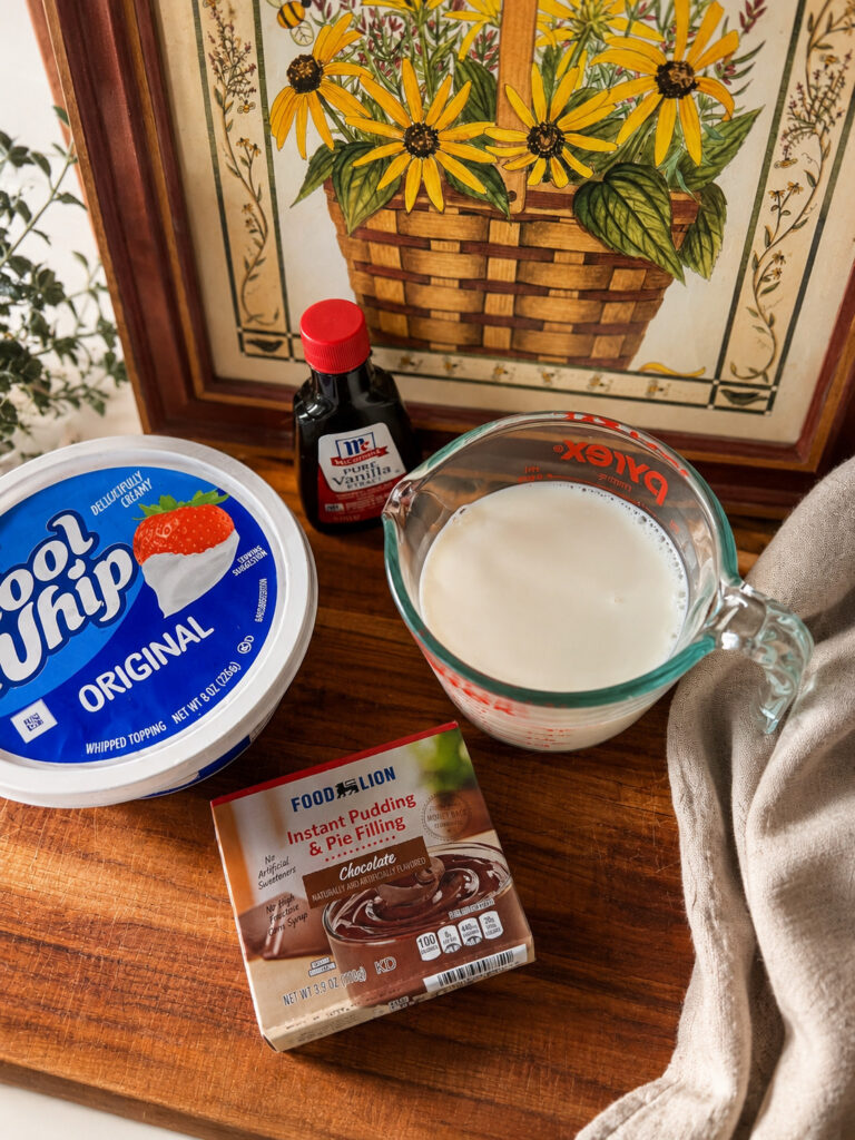 Ingredients for homemade chocolate popsicles including chocolate pudding mix, milk, whipped topping, and vanilla extract arranged on a rustic wooden countertop.