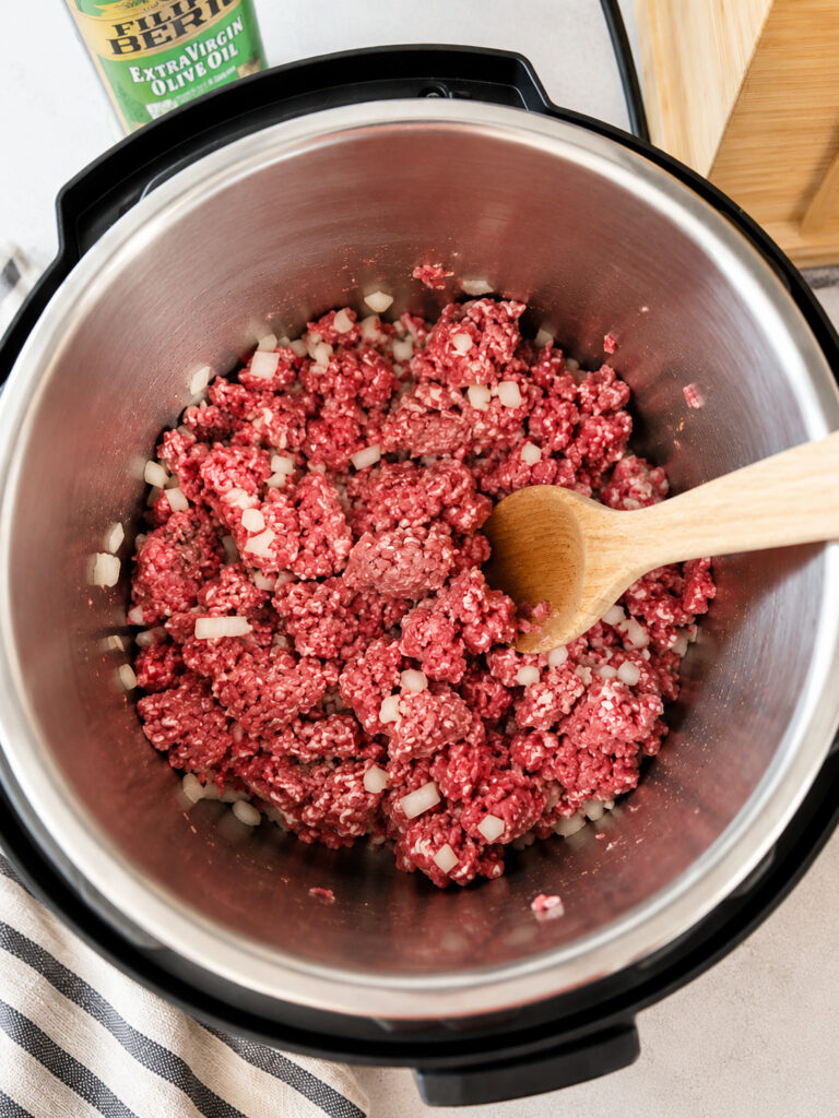 Raw ground beef and diced onion cooking in an Instant Pot with a wooden spoon
