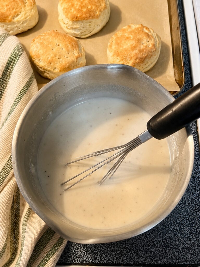 Creamy homemade peppered sausage gravy being whisked in a saucepan with fresh baked biscuits in the background.