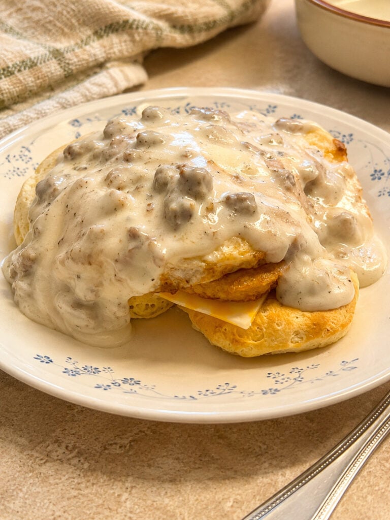 Close-up of loaded biscuits and gravy breakfast sandwich covered in creamy homemade sausage gravy on a vintage plate.