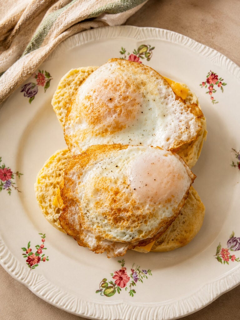 Loaded breakfast biscuit sandwich topped with fried eggs on a floral plate with a striped kitchen towel nearby.