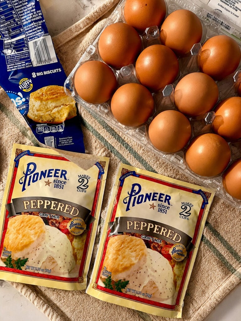 Overhead photo of Pioneer peppered gravy mix packets, eggs in a carton, and canned biscuits arranged on a striped kitchen towel for homemade biscuits and gravy breakfast prep.
