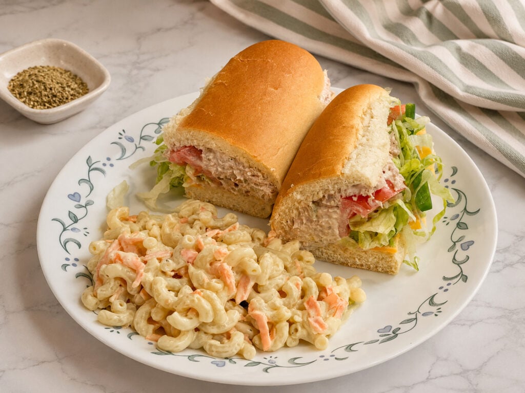 Homemade tuna sandwich served with creamy macaroni salad on a floral plate with shredded lettuce, tomatoes, and soft hoagie bread.