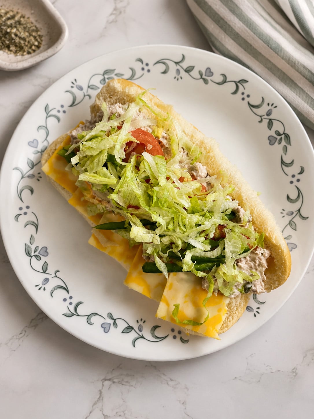 Copycat Subway tuna sandwich recipe on a floral plate with shredded lettuce, tomatoes, green peppers, cheese, and creamy tuna salad on a soft hoagie roll.