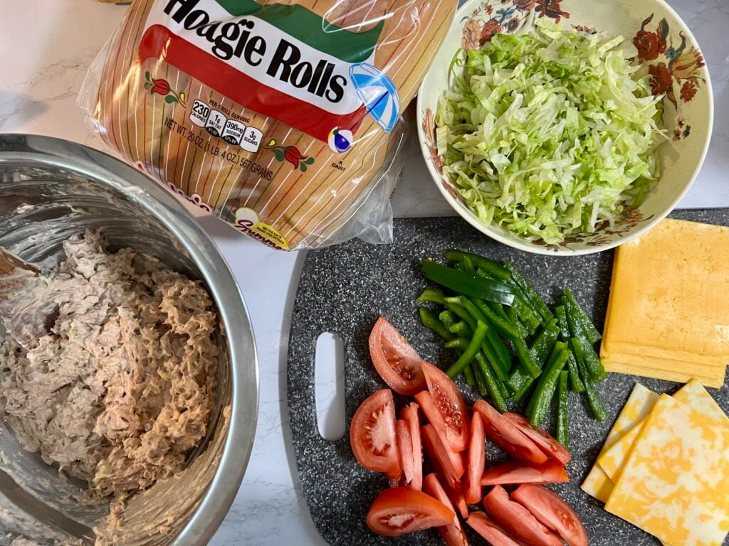 Ingredients for a homemade Subway tuna salad sandwich recipe including hoagie rolls, creamy tuna salad, shredded lettuce, sliced tomatoes, green peppers, and cheese.