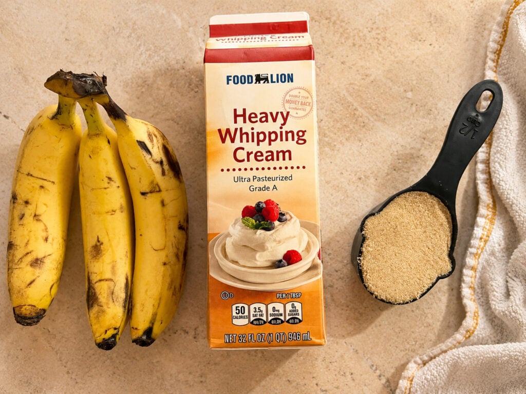 Ingredients for 3 ingredient banana ice cream including ripe bananas, heavy whipping cream, and brown sugar arranged on a countertop with a measuring cup and kitchen towel.