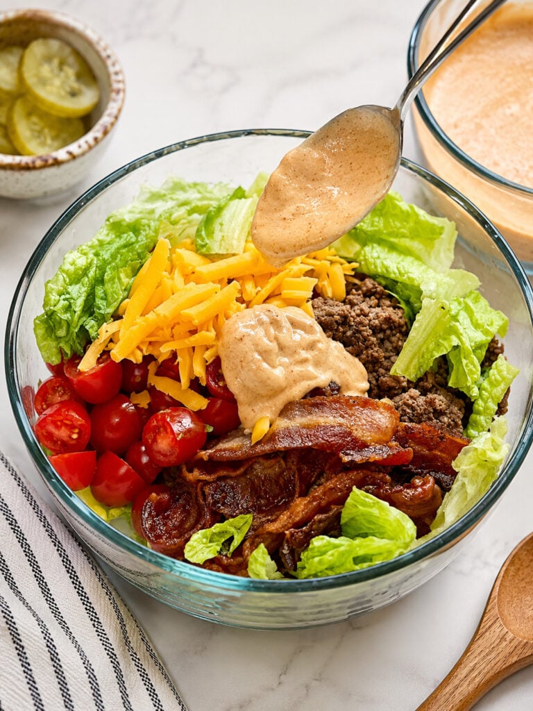 Homemade burger sauce being drizzled over a loaded burger bowl with ground beef, bacon, cheddar cheese, tomatoes, avocado, and romaine lettuce.