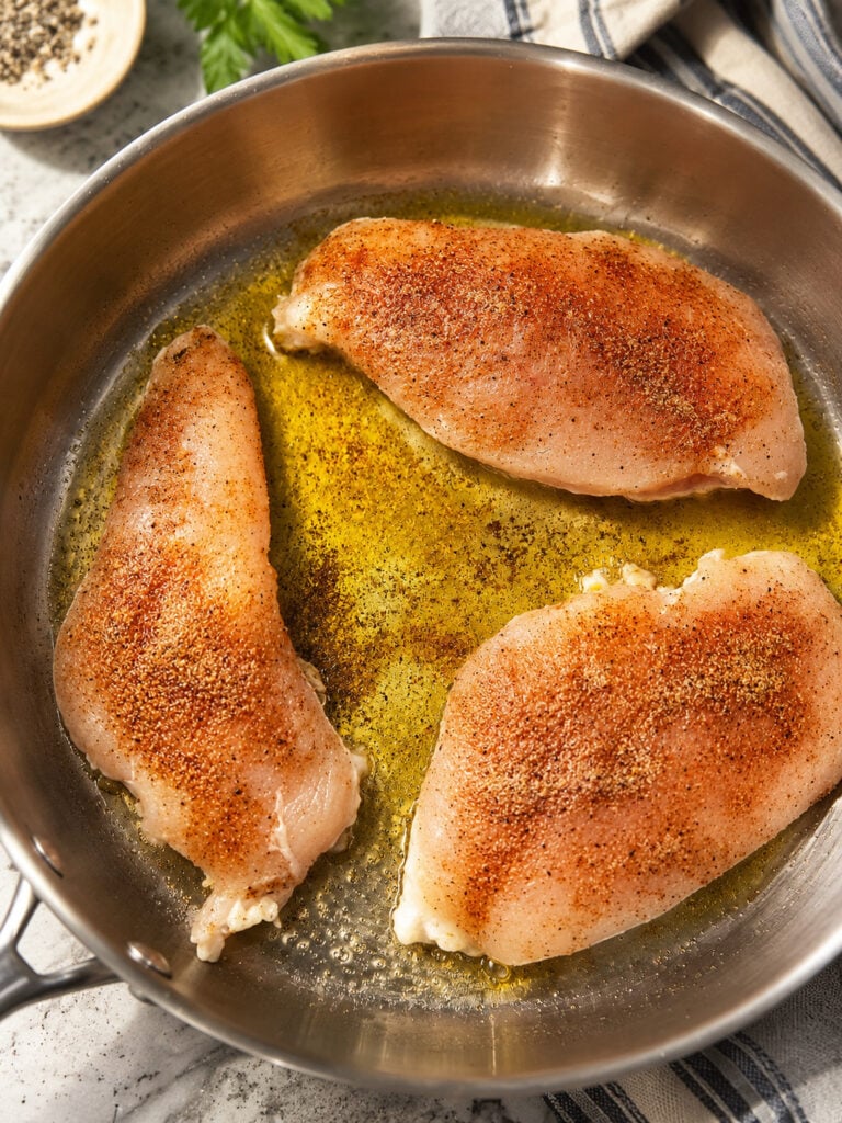 Raw chicken breasts seasoned with paprika, garlic powder, salt, and pepper in a stainless steel skillet with olive oil.