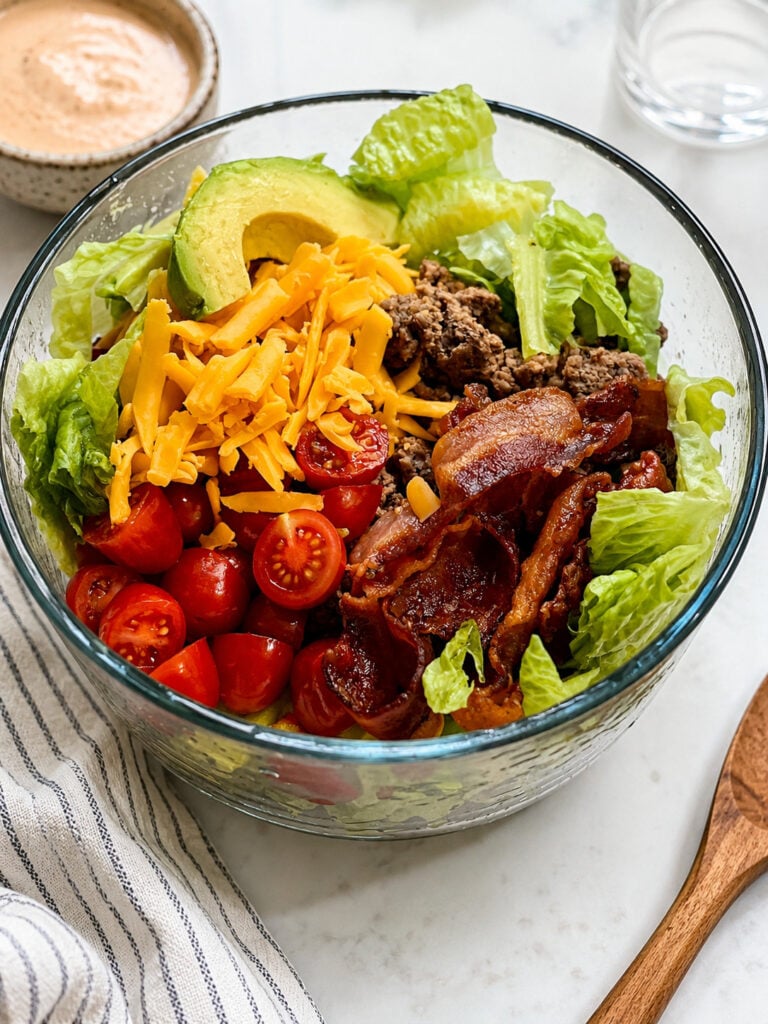 Loaded burger bowl with seasoned ground beef, crispy bacon, cheddar cheese, avocado, cherry tomatoes, romaine lettuce, and homemade burger sauce.