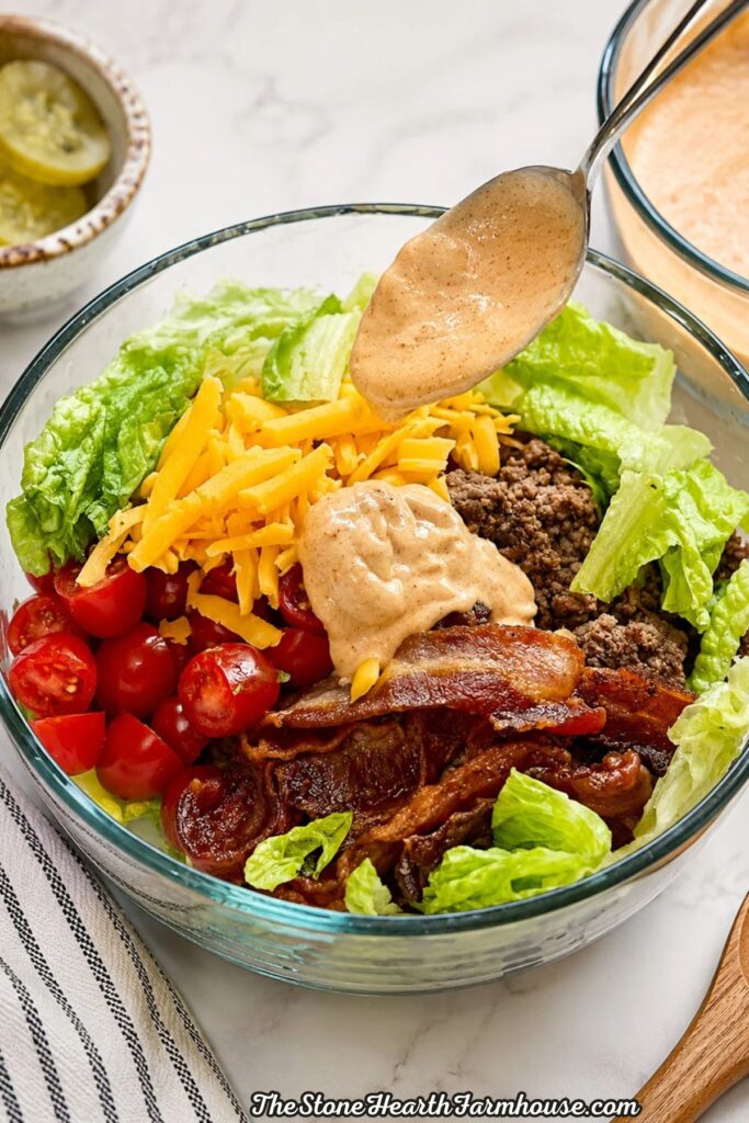 Homemade burger sauce being drizzled over a loaded burger bowl with ground beef, bacon, cheddar cheese, tomatoes, avocado, and romaine lettuce.