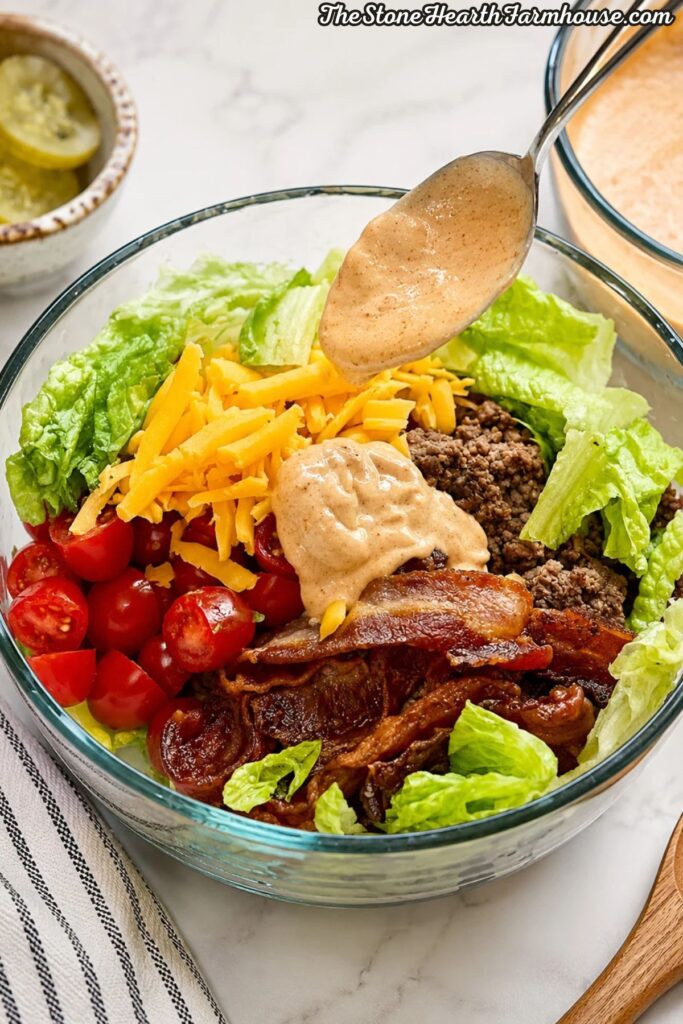 Homemade burger sauce being drizzled over a loaded burger bowl with ground beef, bacon, cheddar cheese, tomatoes, avocado, and romaine lettuce.