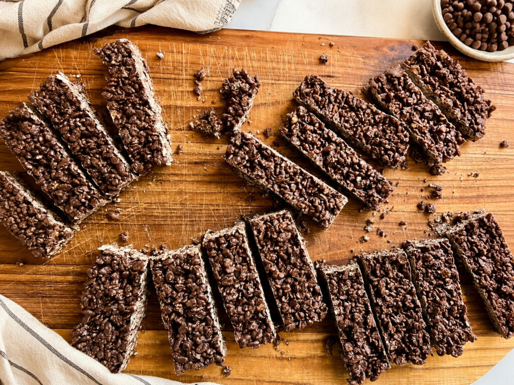 Chocolate Rice Krispie bars sliced into rectangles and arranged on a rustic wooden cutting board with a farmhouse kitchen towel and chocolate chips.