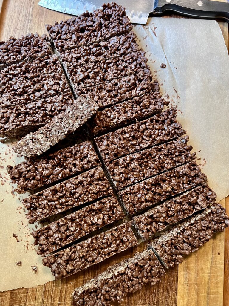 Homemade chocolate Rice Krispie bars sliced into long rectangular bars on parchment paper with a chef's knife.