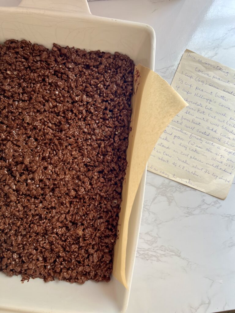 Chocolate Rice Krispie mixture pressed into a parchment-lined baking dish with a vintage handwritten recipe card nearby.