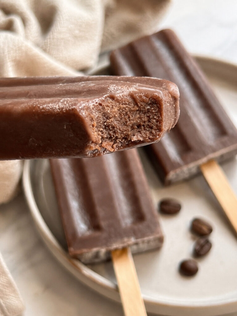 Close-up of a bitten coffee popsicle showing its creamy frozen chocolate coffee pudding texture with additional coffee popsicles in the background.