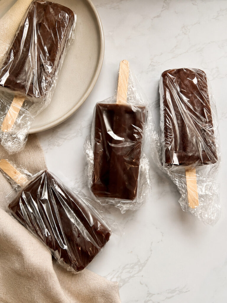Frozen coffee popsicles individually wrapped in plastic wrap for freezer storage on a marble countertop.