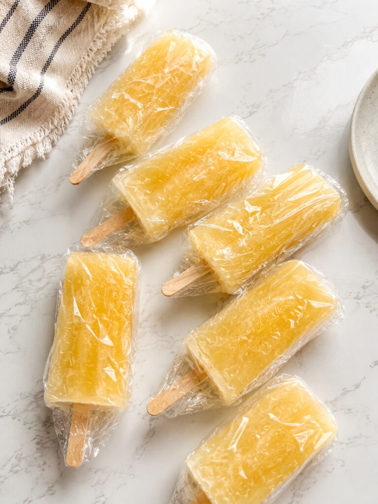 Homemade pineapple popsicles individually wrapped in plastic wrap for freezer storage on a marble countertop.