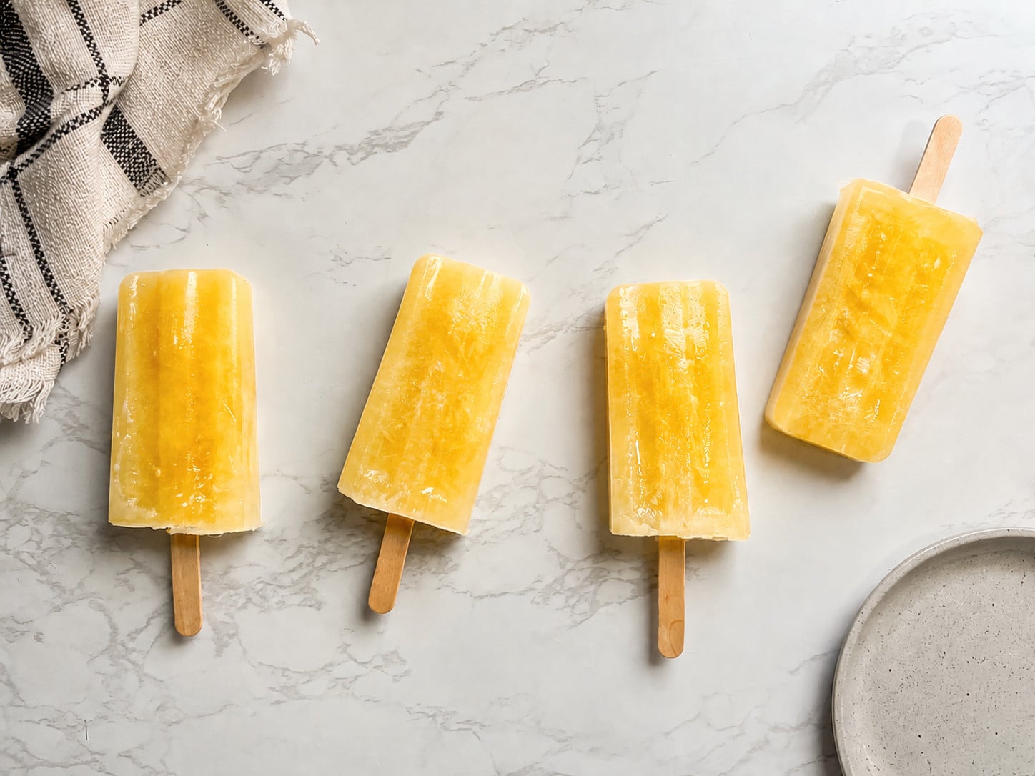 Homemade pineapple popsicles made with 100% pineapple juice arranged on a marble countertop with a kitchen towel and ceramic plate.