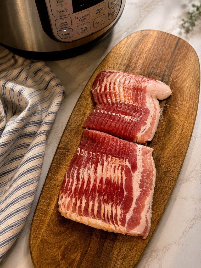 Uncooked bacon slices arranged on a wooden serving board beside an Instant Pot and striped kitchen towel.