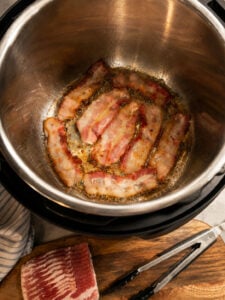 Bacon strips sizzling in the Instant Pot using the sauté function with rendered bacon grease forming in the bottom.