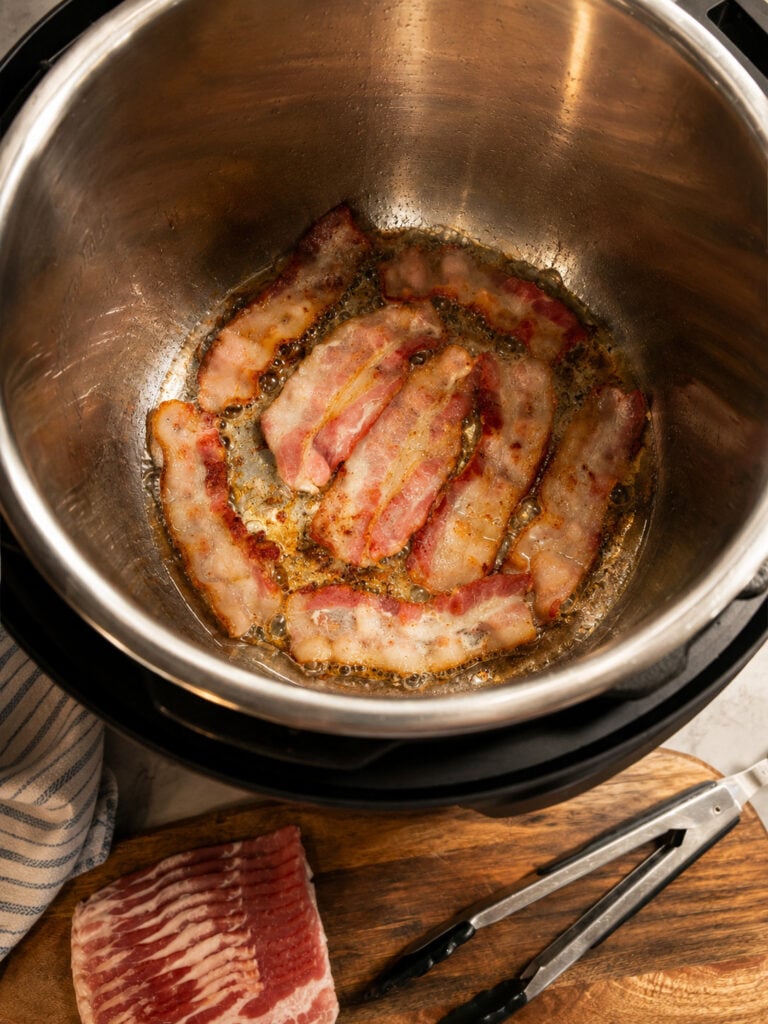 Bacon strips sizzling in the Instant Pot using the sauté function with rendered bacon grease forming in the bottom.