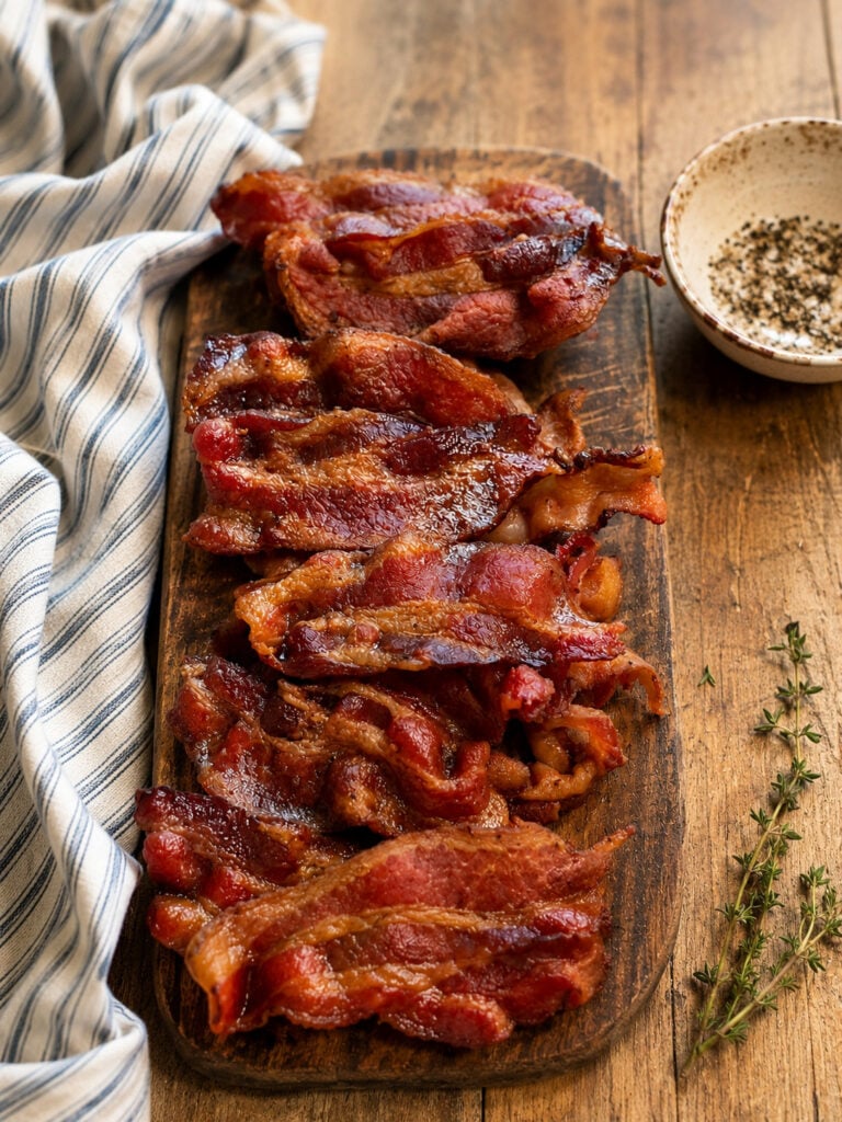 Perfectly cooked crispy bacon piled on a wooden board with a rustic kitchen towel and seasoning dish.