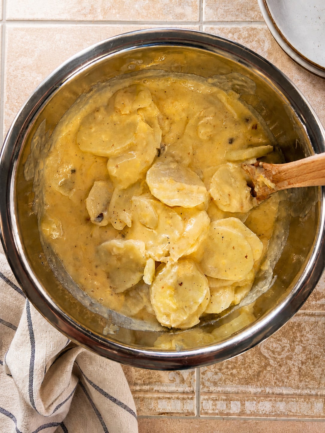 **Alt text:** Overhead view of creamy Instant Pot scalloped potatoes in a stainless steel mixing bowl with a wooden spoon. Thinly sliced potatoes are coated in a rich cheese sauce made with cream, sour cream, and cheese. A beige striped kitchen towel sits beside the bowl on a vintage-style tiled countertop.