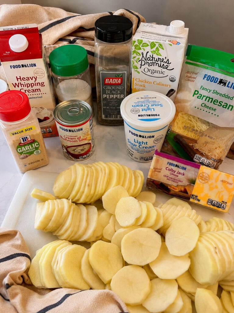 Ingredients for Instant Pot scalloped potatoes including sliced potatoes, heavy cream, sour cream, cream of mushroom soup, chicken stock, garlic powder, black pepper, Parmesan cheese, and Colby Jack cheese arranged on a countertop.