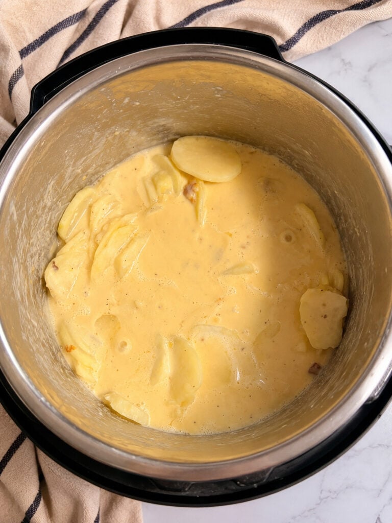 Thinly sliced potatoes coated in a creamy cheese sauce inside an Instant Pot insert before pressure cooking for scalloped potatoes.