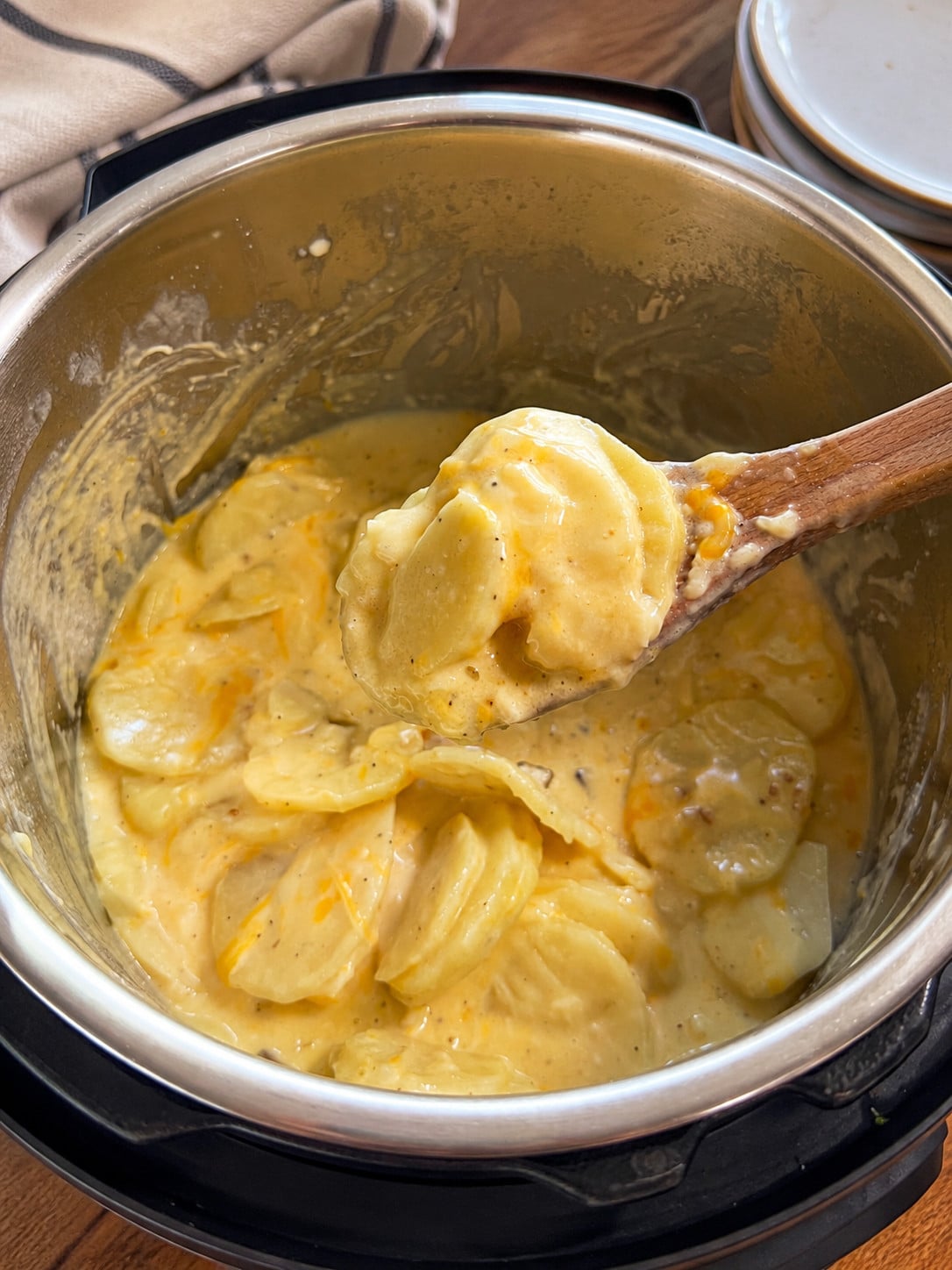 Creamy Instant Pot scalloped potatoes coated in a rich cheesy sauce, served from a wooden spoon over the Instant Pot insert with a beige striped kitchen towel in the background.