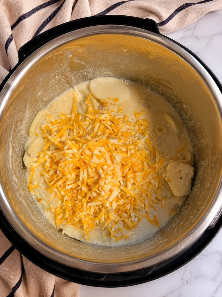 Shredded Colby Jack and Parmesan cheese sprinkled over sliced potatoes in a creamy sauce inside an Instant Pot for cheesy scalloped potatoes.