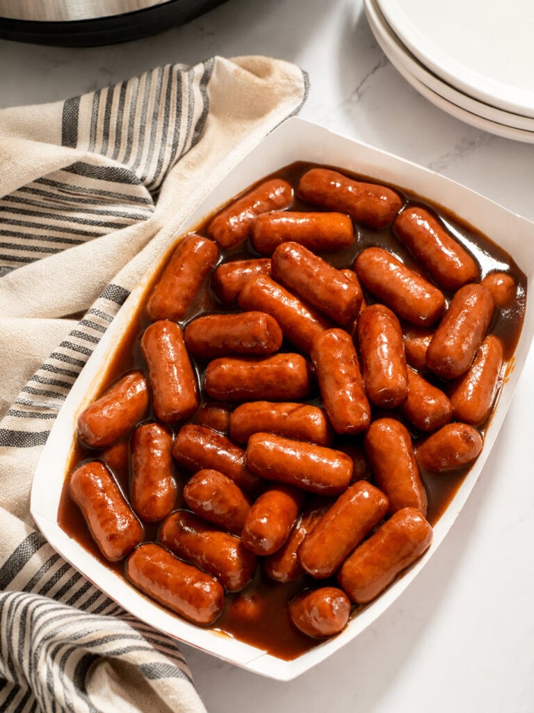 Instant Pot BBQ Little Smokies served in a white paper tray with sweet barbecue sauce, styled with a striped kitchen towel and white plates on a marble countertop.
