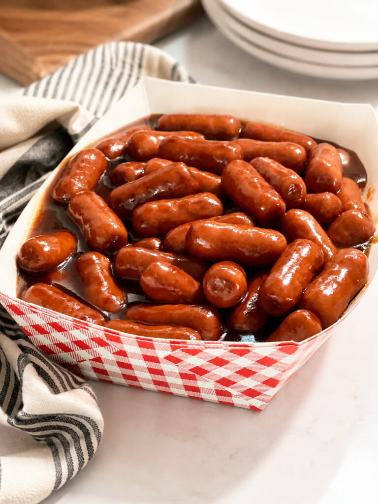 Close-up of BBQ Little Smokies appetizer served warm in a paper tray with rich barbecue sauce and a striped kitchen towel.