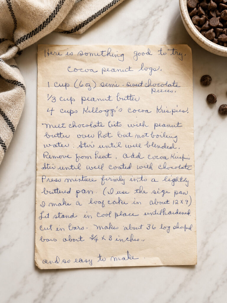 Handwritten vintage recipe card for Cocoa Peanut Logs displayed on a marble countertop with chocolate chips and a farmhouse towel.