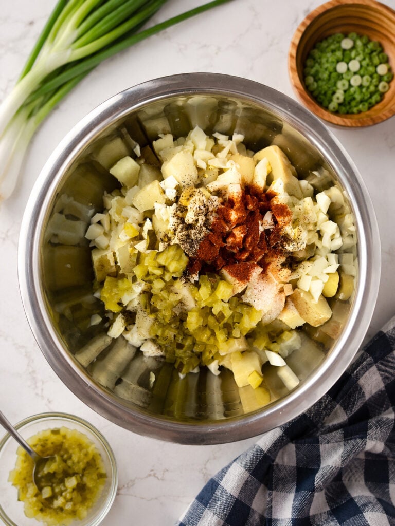 Potatoes, chopped eggs, dill pickles, mayonnaise, mustard, and seasonings combined in a mixing bowl before stirring into a creamy side dish.