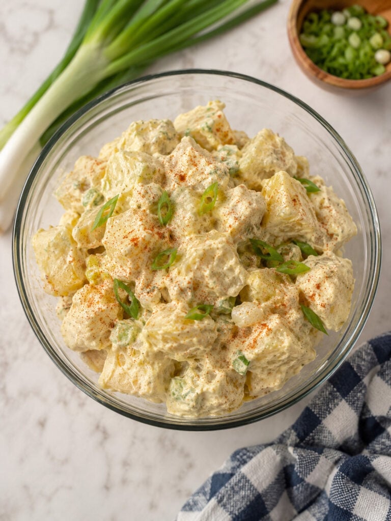 Creamy homemade potato salad with hard-boiled eggs, dill pickles, green onions, and paprika served in a glass bowl for a classic summer side dish.