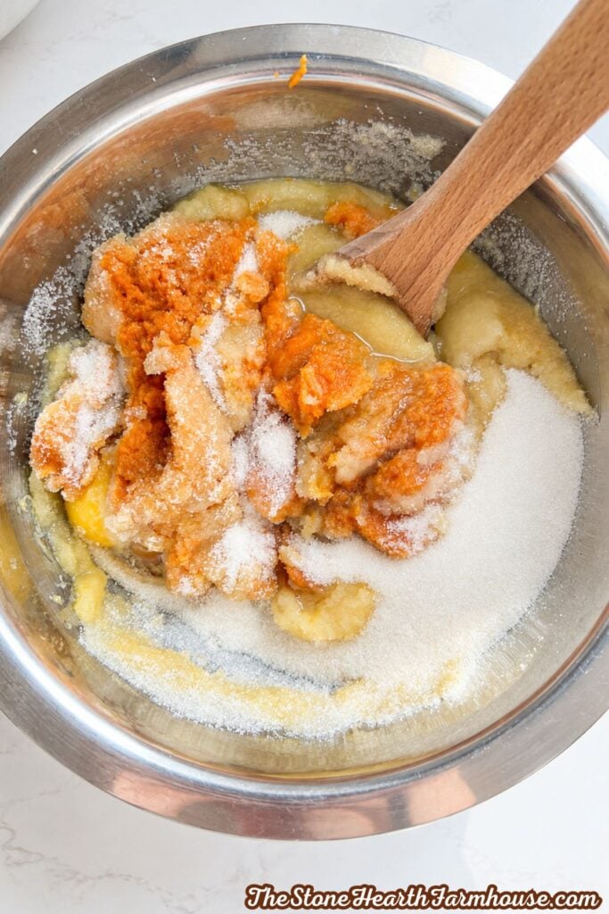Pumpkin purée mixture and granulated sugar being combined in a stainless steel bowl with a wooden spoon