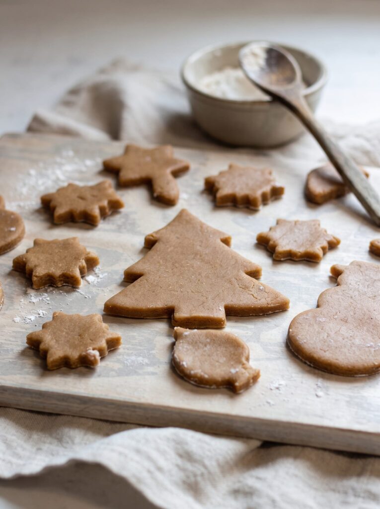Unbaked ginger spice sugar cookies cut into tree, star and snowman shapes on a floured wooden board beside a bowl of flour